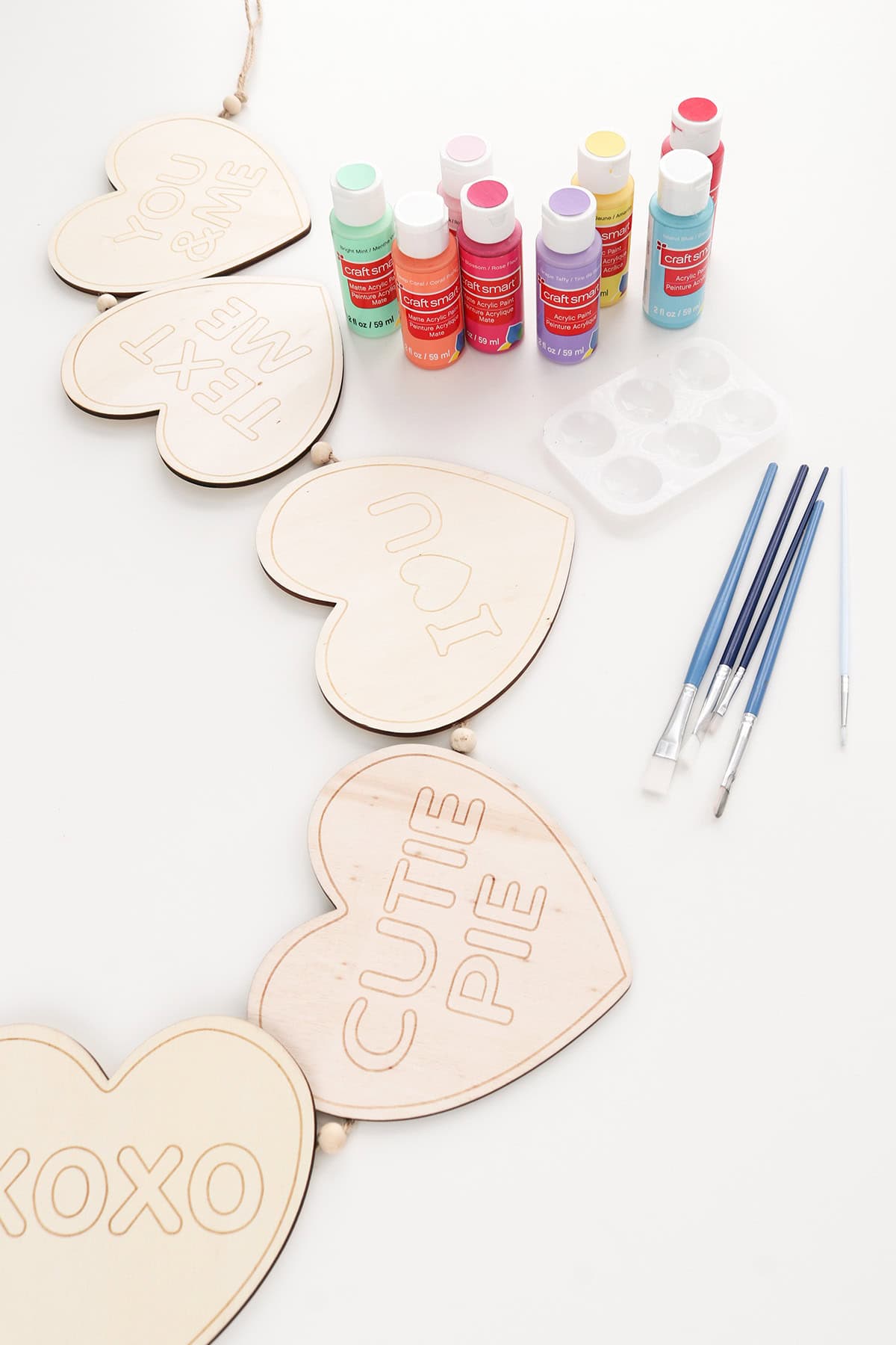 supplies needed for a diy wood conversation heart wreath, unfinished wood heart garland and bottles of paint with brushes on white tabletop