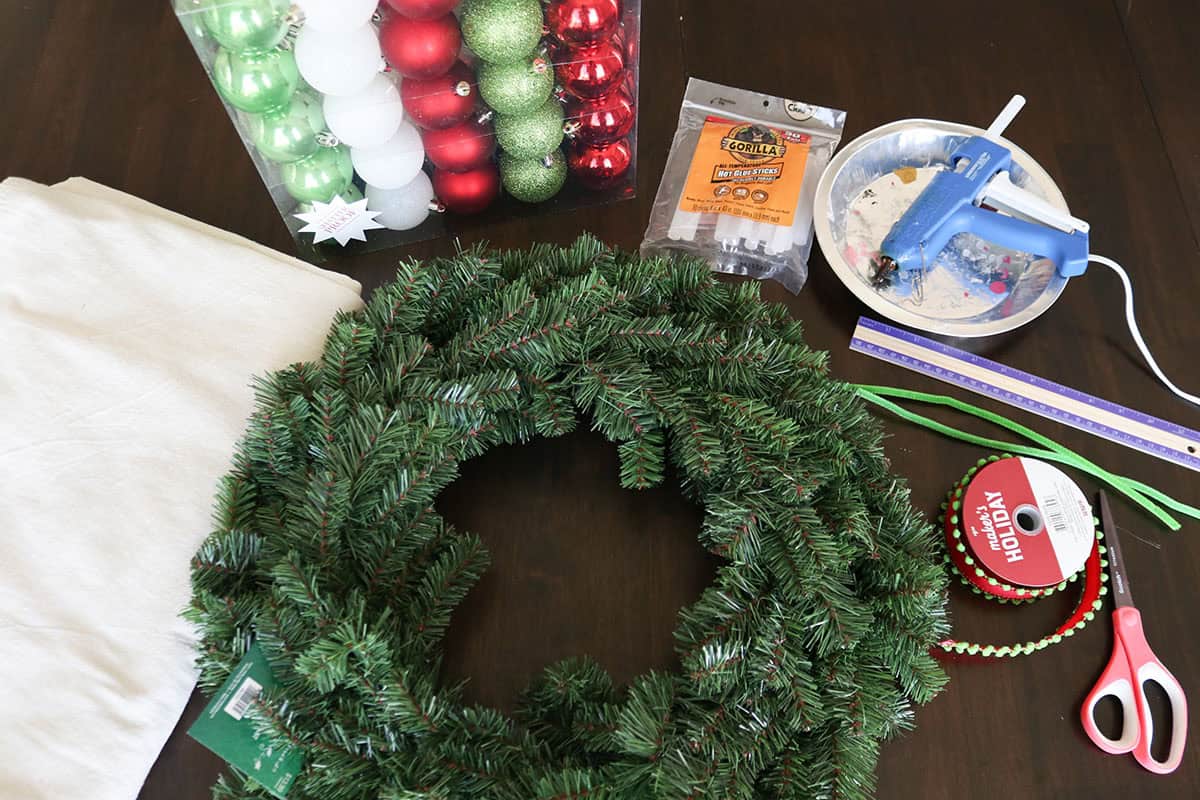 supplies to make a diy evergreen Christmas ornament wreath including wreath form, ornaments, hot glue, and ribbon